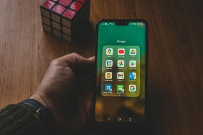 A hand holds a smartphone displaying various apps, next to a Rubik's cube on a wooden surface.
